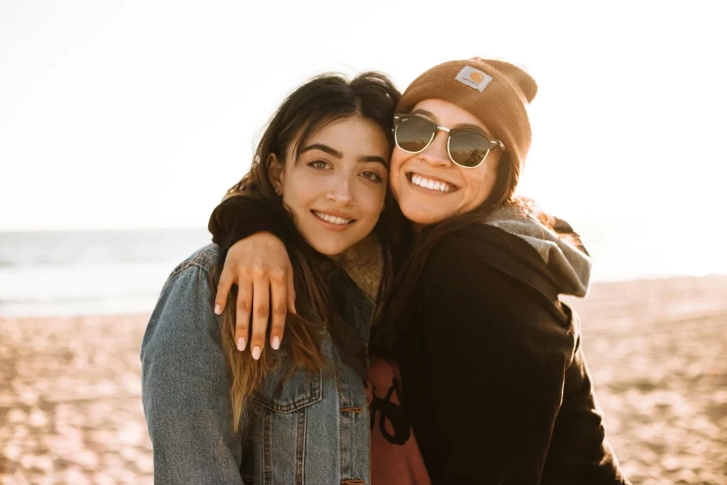Dos personas abrazándose en una playa de arena con el océano de fondo al atardecer. Una lleva una chaqueta vaquera y la otra una sudadera negra con capucha y gafas de sol.